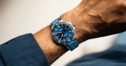 A blue dive watch with a rubber strap on a persons wrist, wearing a blue long-sleeve shirt.