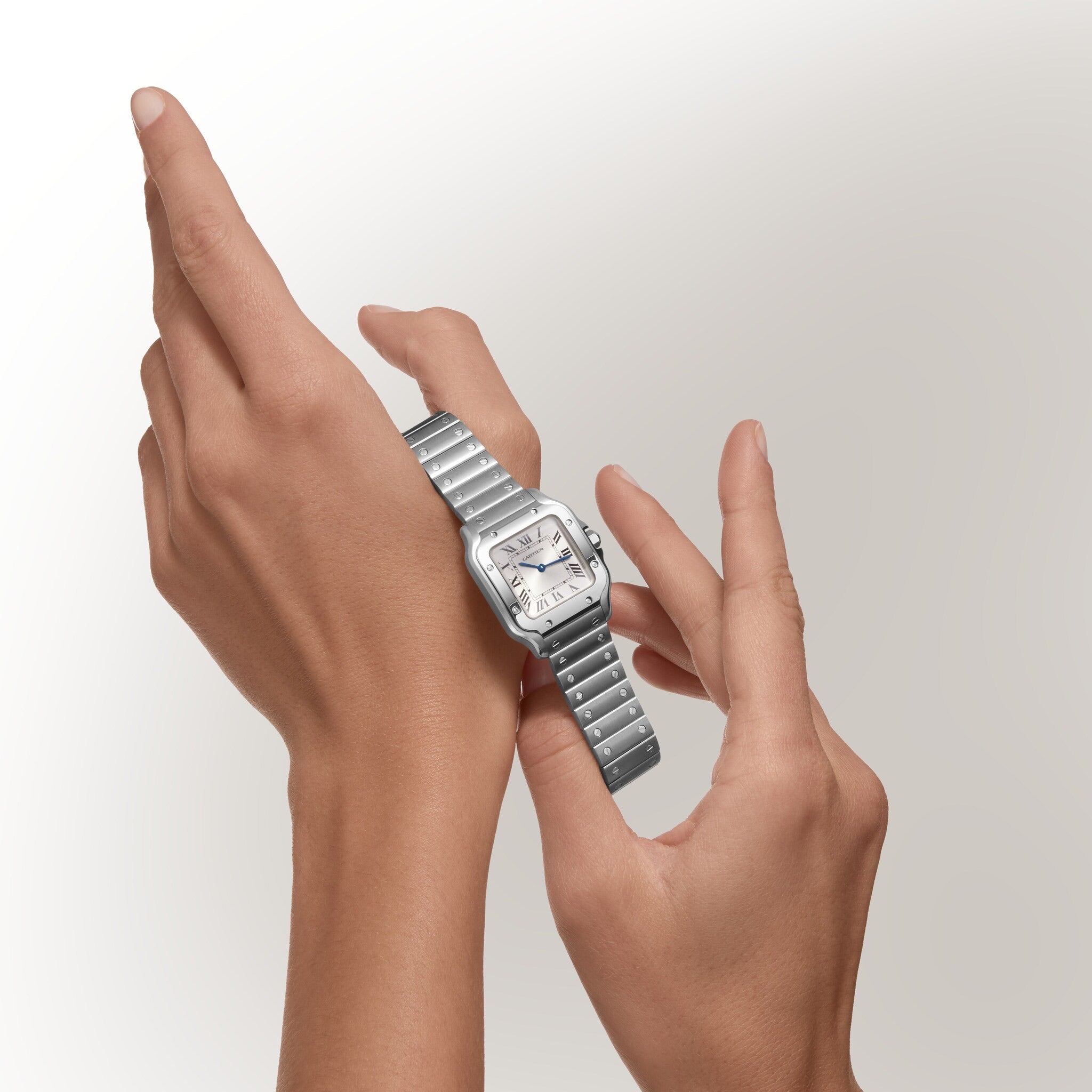 A person adjusts a Santos De Cartier silver wristwatch by Cartier on their wrist against a light, plain background.