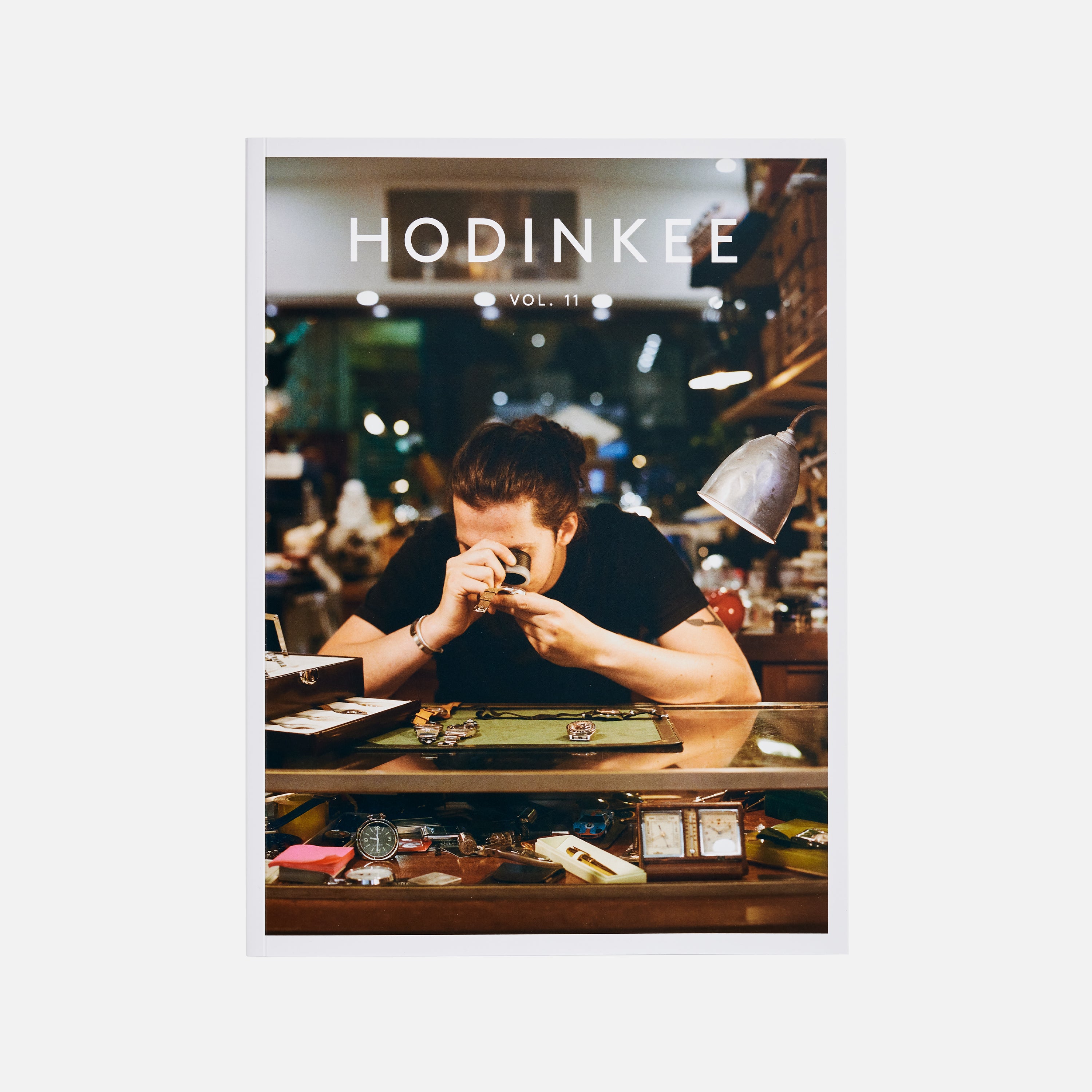 A person inspects a watch at a cluttered counter under a lamp, beside HODINKEE Magazine, Volume 11 by Hodinkee, showcasing the print journal’s passion for Bauhaus design and fine watchmaking.