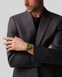 A man in a dark suit crosses his arms against a light gray background, wearing the Zenith Defy Revival black and yellow watch.
