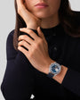 Woman in a black top rests her face on her hand, showcasing the Zenith Chronomaster Original—a striking blue and silver wristwatch.