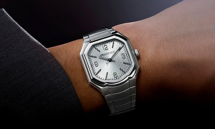 A wrist with a dark gray shirt sleeve and a Gerald Charles Masterlink watch with a silver-toned strap and dial