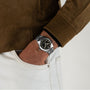 A person in a brown suede jacket and white pants keeps their left hand in their pocket, revealing a vintage-style Hamilton Khaki Field Murph 38mm watch with a black dial and Arabic numerals.