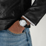 A person in a black leather jacket and blue jeans has their left hand in their pocket, showcasing the Hamilton Khaki Field Murph 38mm—a vintage-inspired timepiece with a white face and black leather strap beloved by enthusiasts.