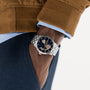 A person in a brown jacket and blue pants wears the Hamilton Jazzmaster Open Heart Auto 42mm, displaying its dark openwork dial and automatic movement, with their hand in their pocket.