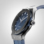 A close-up of the Hublot Classic Fusion Ceramic Blue wristwatch with a black ceramic bezel, silver markers, blue leather strap, and HUB1112 automatic movement, photographed against a light gradient background.