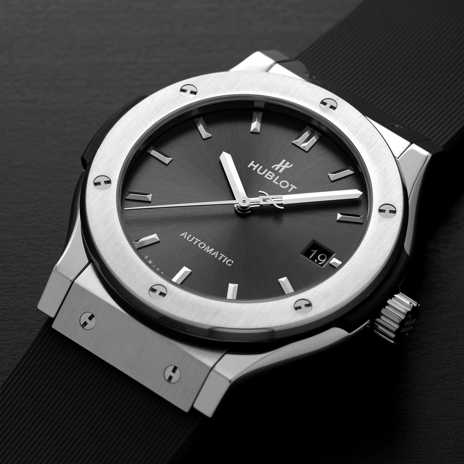 A close-up of the Hublot Classic Fusion Racing Grey Titanium wristwatch, featuring a grey dial, silver markers, date display, black rubber strap, and a sleek titanium case on a dark surface.