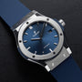 The Hublot Classic Fusion Titanium Blue features a blue dial and strap, silver bezel, and automatic movement, displayed on a black surface.