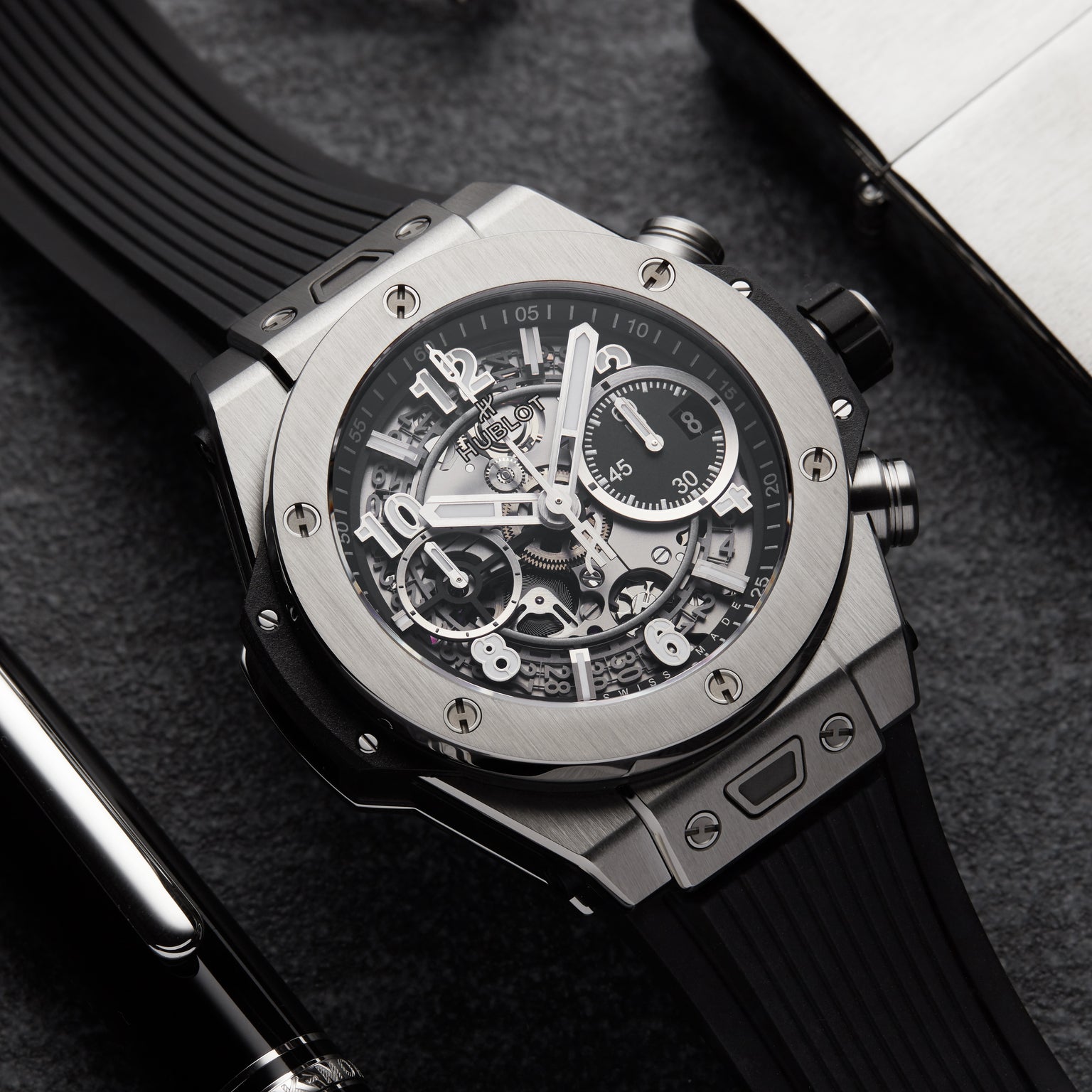 A close-up of the Hublot Big Bang Unico Titanium, highlighting its skeleton chronograph flyback movement, black rubber strap, and titanium case on a dark surface beside a pen and metallic object.