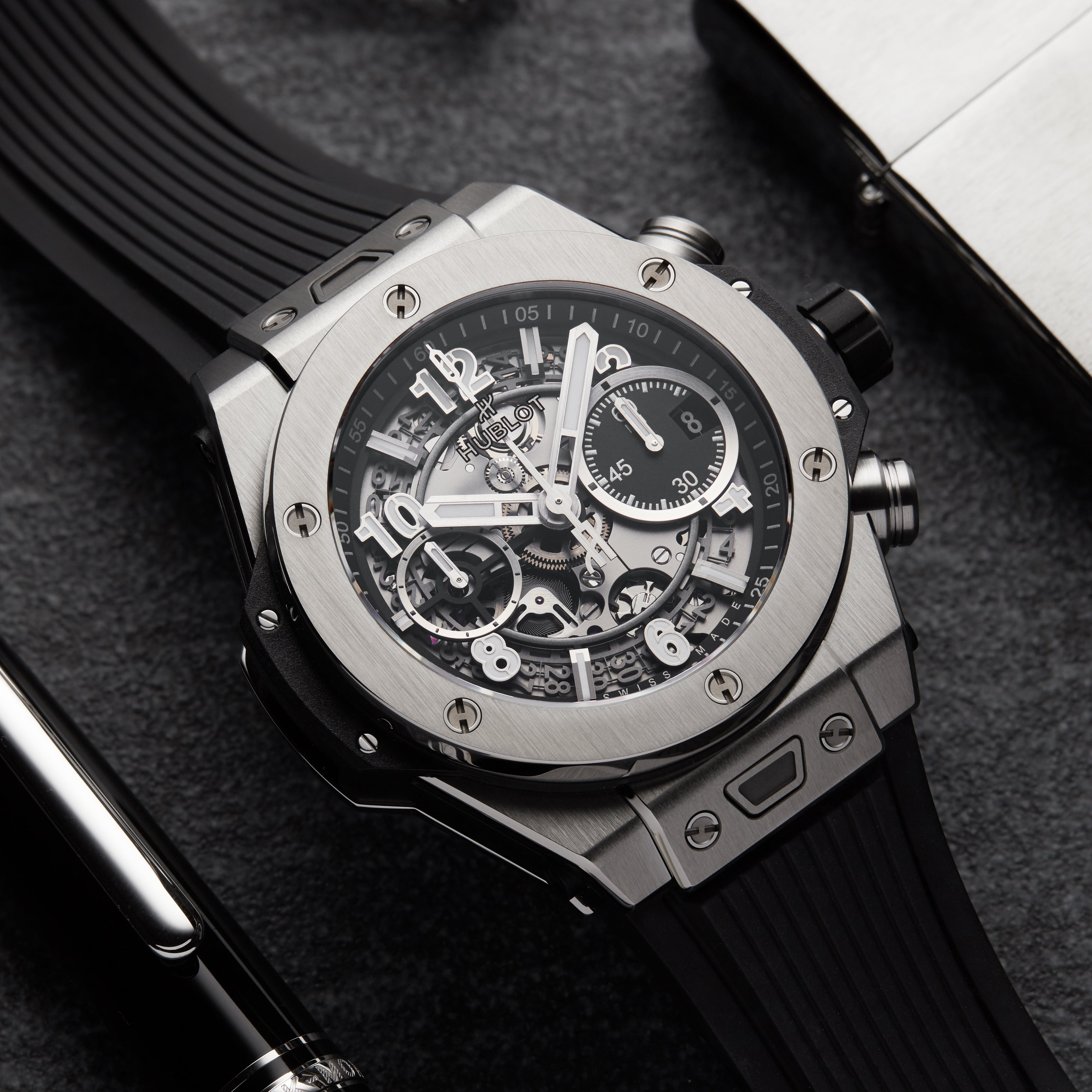 A close-up of the Hublot Big Bang Unico Titanium, highlighting its skeleton chronograph flyback movement, black rubber strap, and titanium case on a dark surface beside a pen and metallic object.