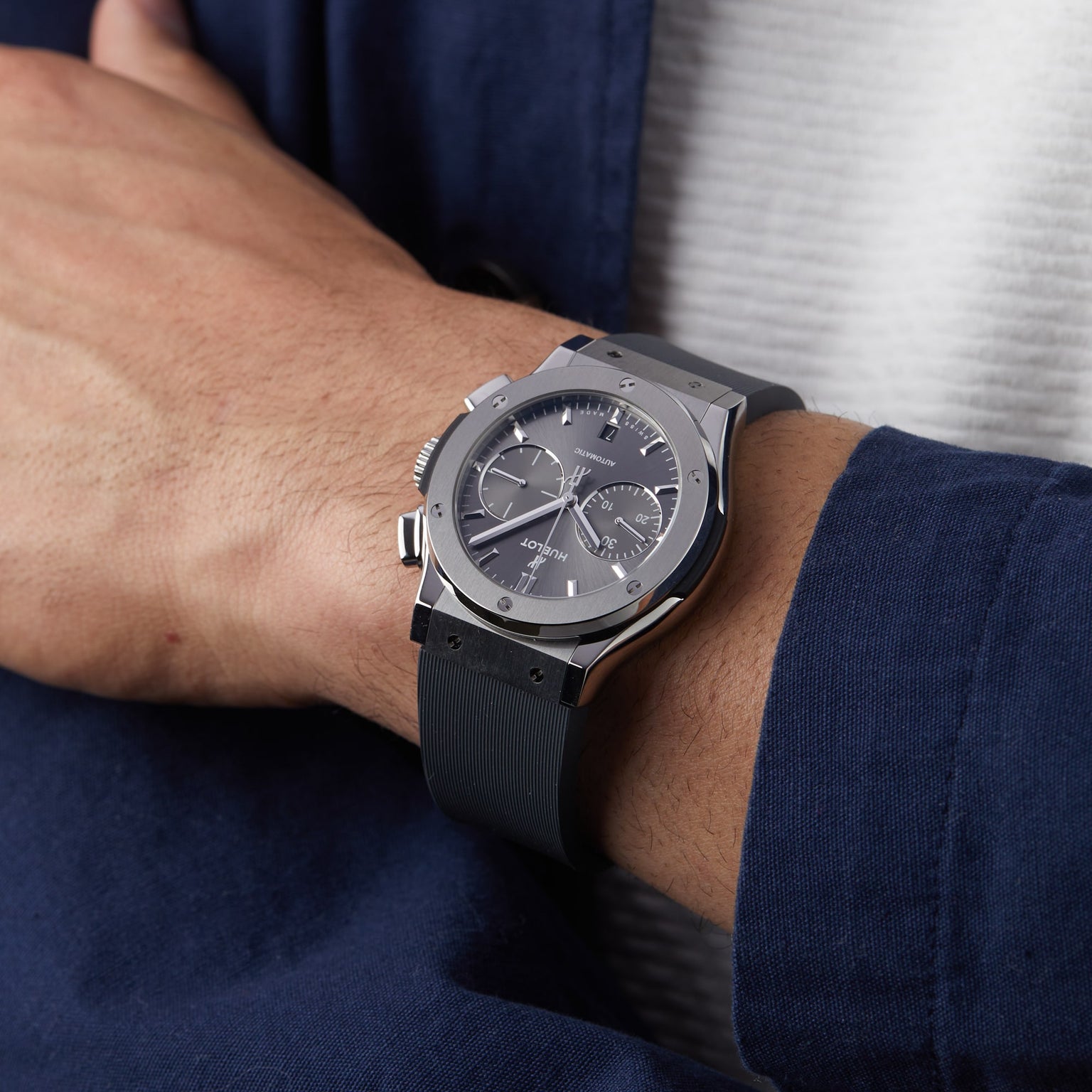 A person wearing a Hublot Classic Fusion Racing Grey Chronograph Titanium watch with a black strap and a dark blue jacket over a white shirt.