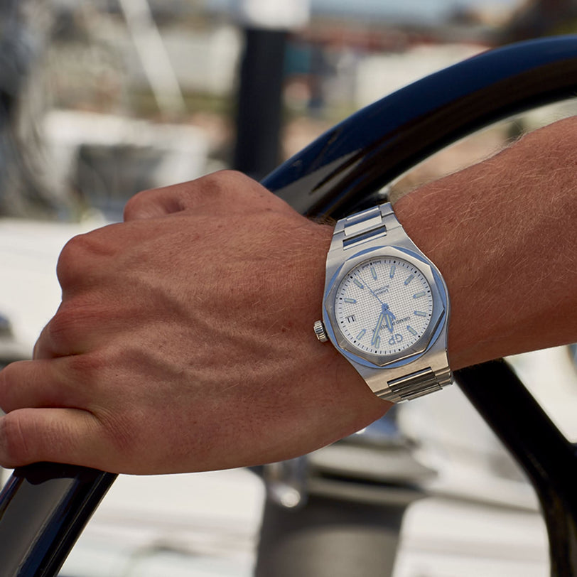 A wrist wearing the Girard-Perregaux Laureato 42, with its octagonal bezel and stainless steel case, grips a black steering wheel against a blurred outdoor background in daylight.