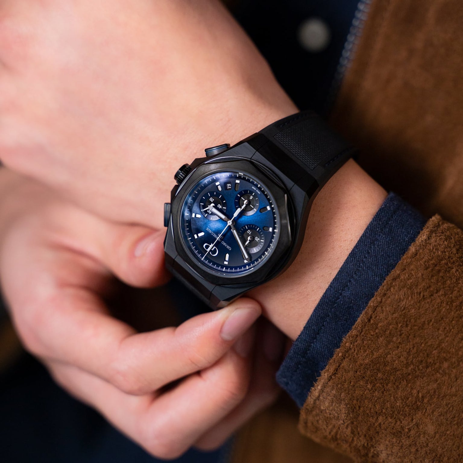 A person in a brown jacket checks a Girard-Perregaux Laureato Absolute Chronograph with a black PVD-treated titanium case and black strap on their left wrist.