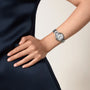 A person in a sleeveless dark dress rests their hand on their hip, showcasing the Cartier Ballon Bleu de Cartier watch with its round face and signature sapphire cabochon.