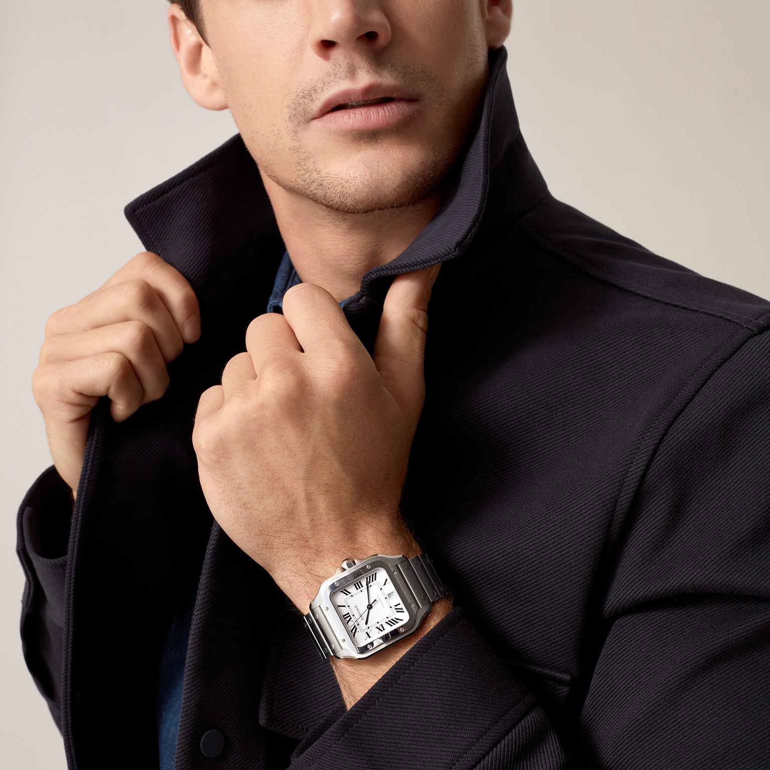 A man in a dark jacket adjusts his collar, showcasing a Cartier Santos de Cartier silver wristwatch with a square face on his wrist.