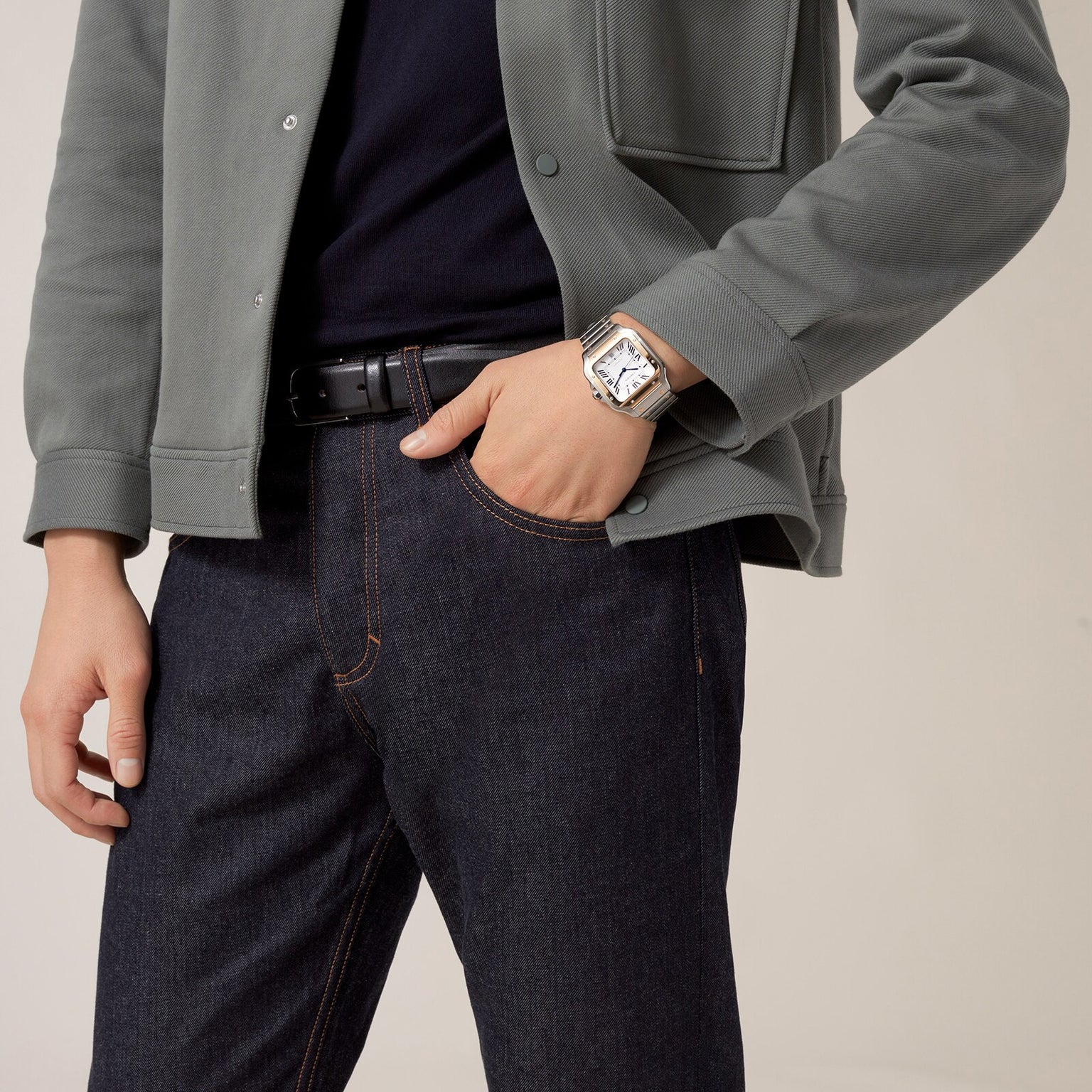 A person in a green jacket, black shirt, and dark jeans wears a Cartier Santos de Cartier silver wristwatch with their hand in their pocket, standing before a plain background—a minimal and timeless statement.