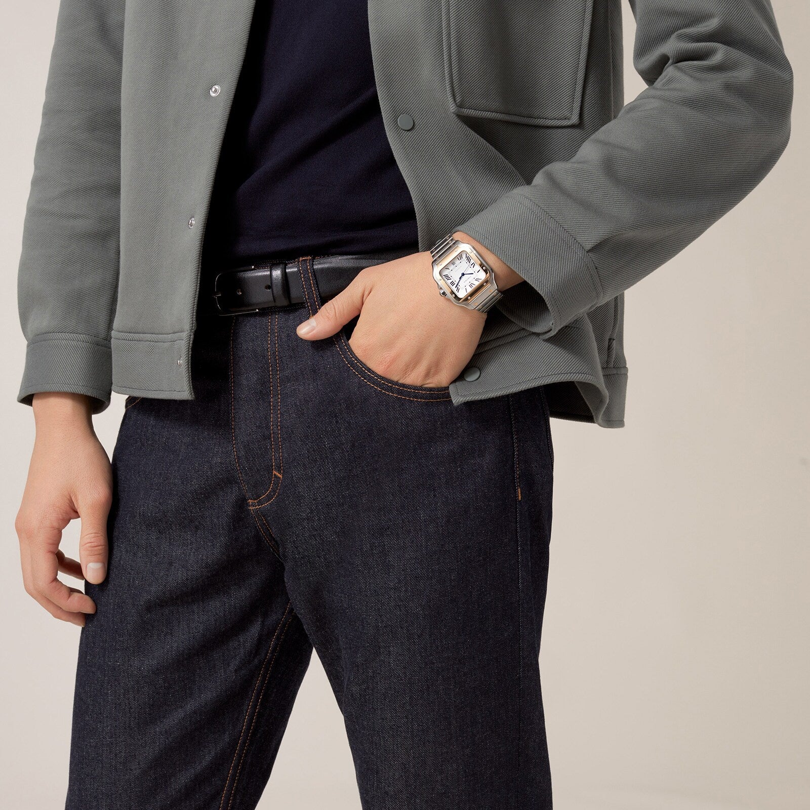 A person in a green jacket, black shirt, and dark jeans wears a Cartier Santos de Cartier silver wristwatch with their hand in their pocket, standing before a plain background—a minimal and timeless statement.