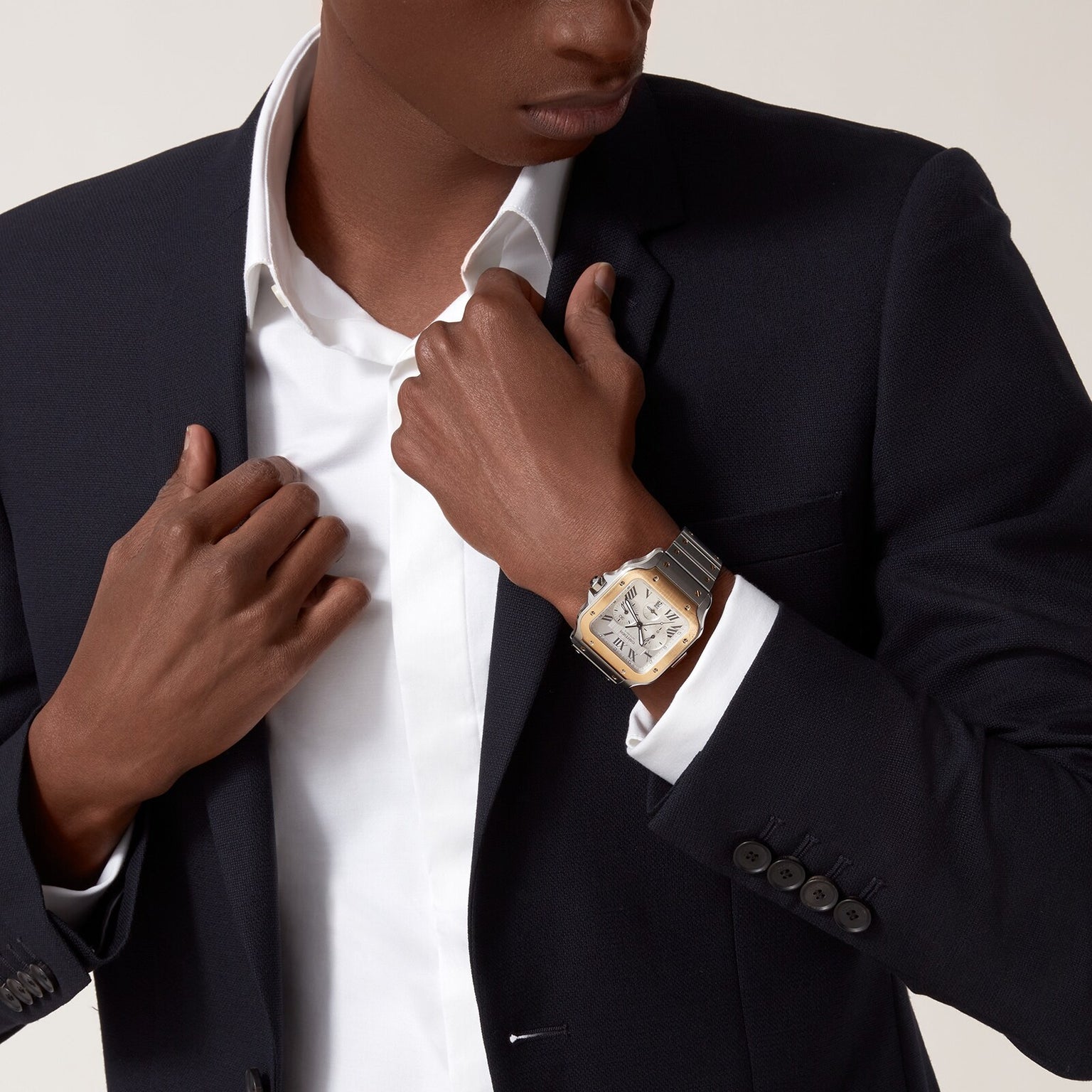 A person in a dark suit and white shirt adjusts their jacket, revealing a Cartier Santos de Cartier Chronograph watch with a metallic band on their wrist.