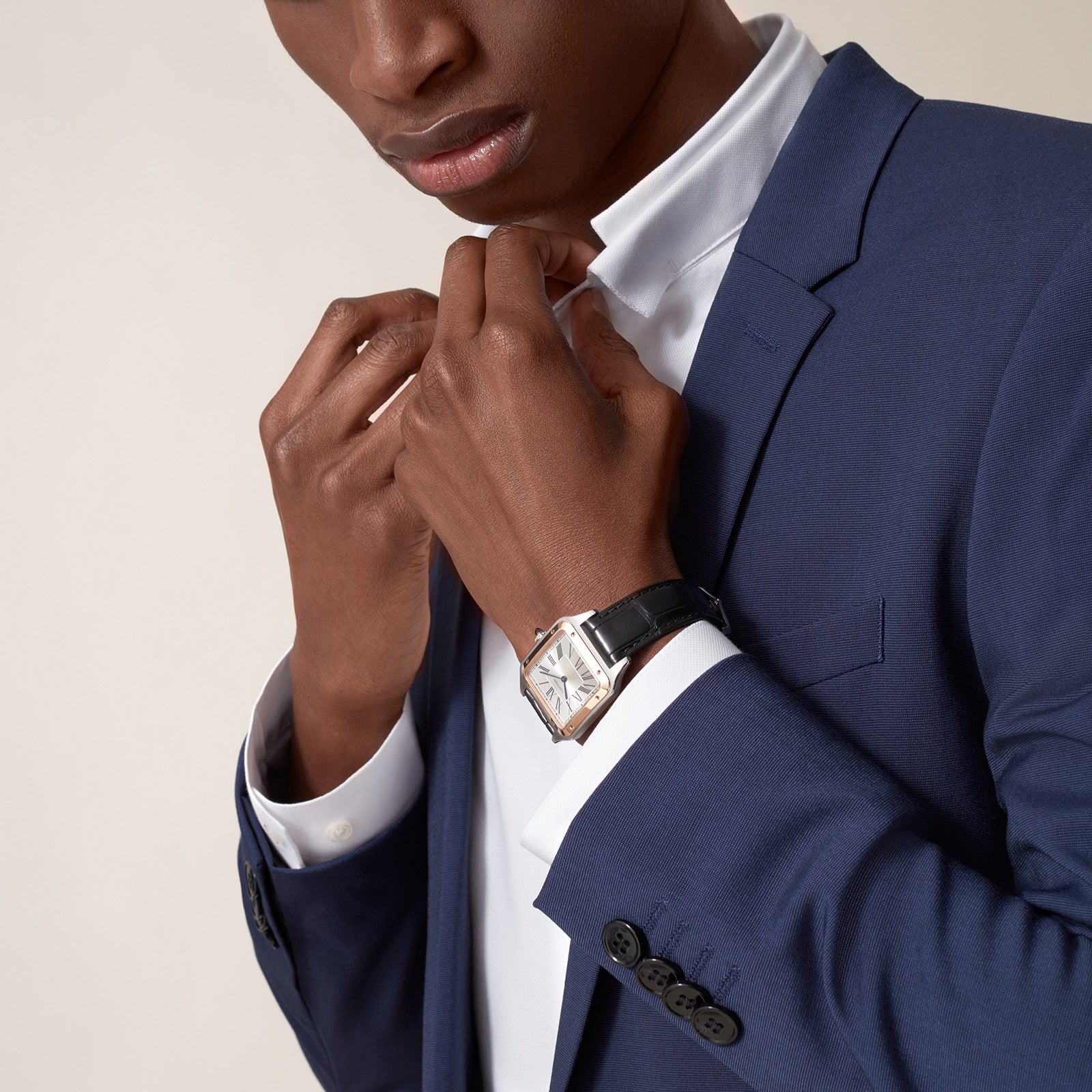 A person in a blue suit adjusts the collar of a white shirt, wearing a Ballon Bleu de Cartier watch by Cartier with a black leather strap on their wrist.
