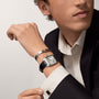 A suited individual displays a wrist adorned with a Cartier Santos-Dumont watch featuring a rectangular face, black leather strap, and silver bracelet.