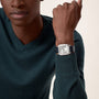 A person in a dark green sweater showcases the Cartier Santos-Dumont watch, which has a square face, mechanical movement, Roman numeral markers, and a black leather strap.