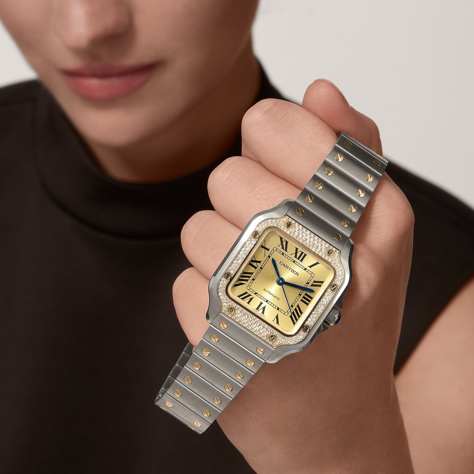 A person wearing a sleeveless dark top raises their wrist to reveal the Cartier Santos de Cartier Chronograph, featuring a gold and silver design, square face, Roman numeral markers, and a diamond bezel.