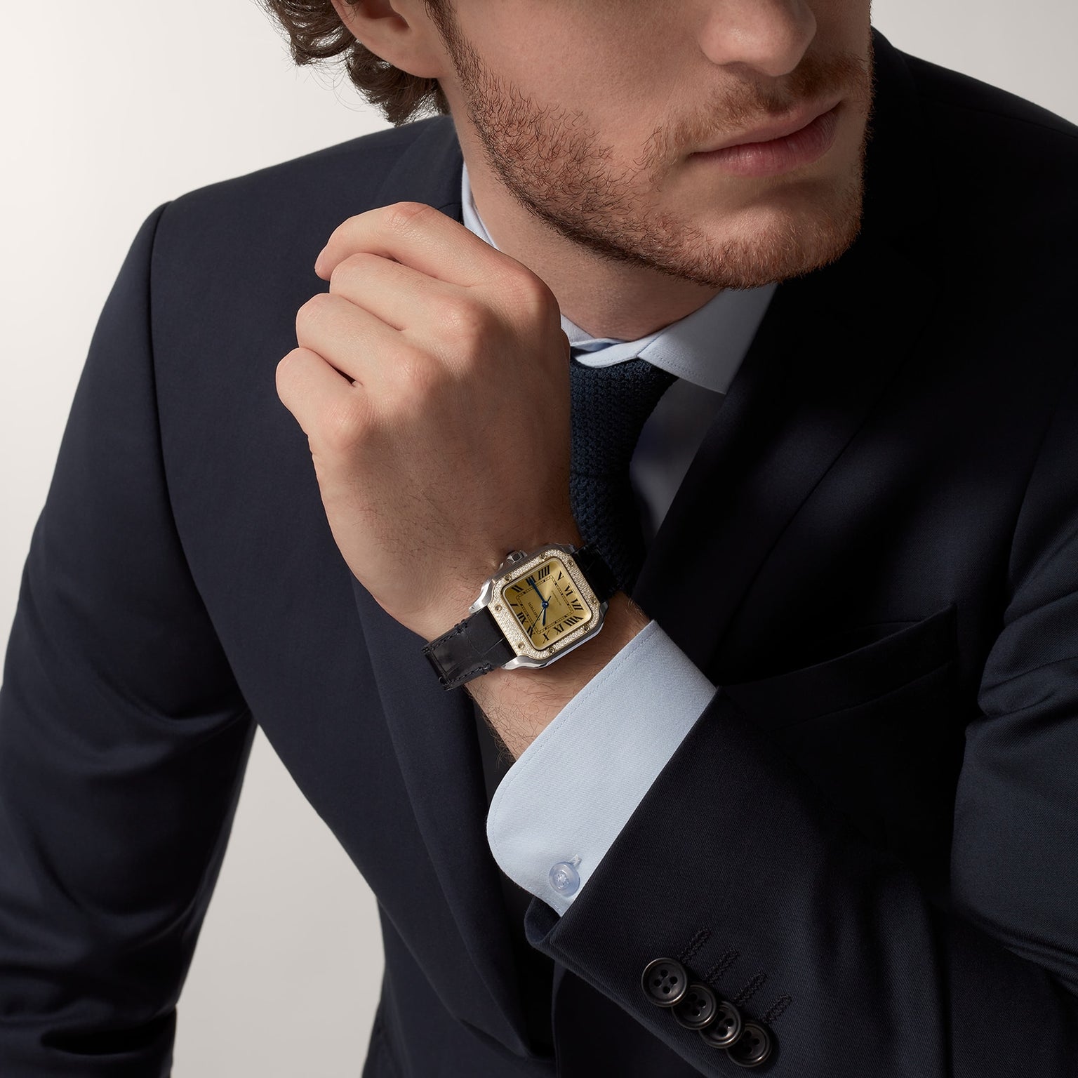 A man in a dark suit and tie adjusts his cuff, revealing a Cartier Santos de Cartier Chronograph with a gold dial, black leather strap, and mechanical movement.