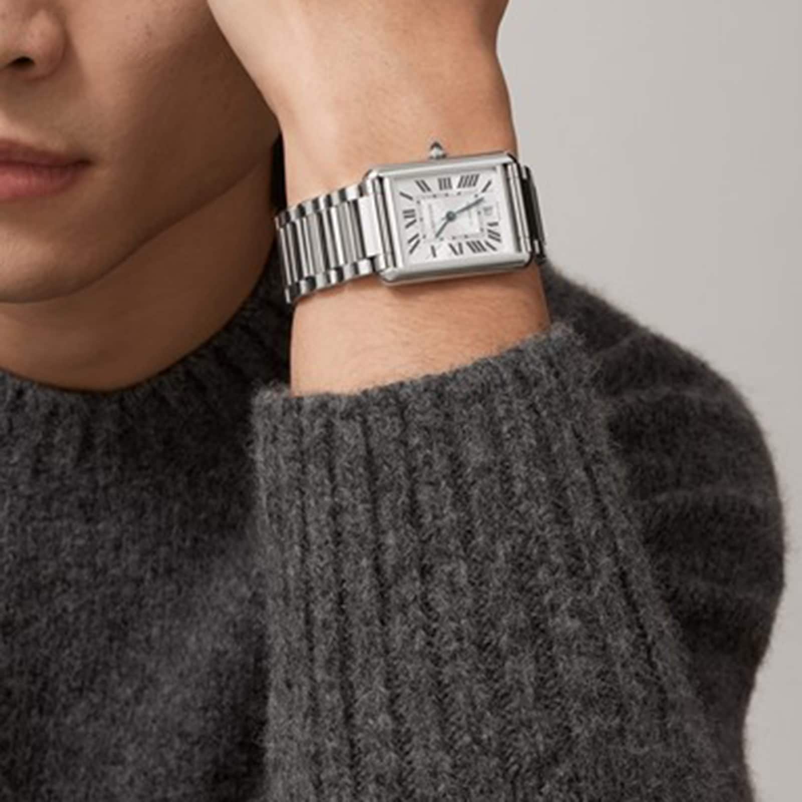 A person in a grey knit sweater showcases the Cartier Tank Américaine watch, featuring a rectangular silvered flinqué dial with Roman numeral markers on a metal band.