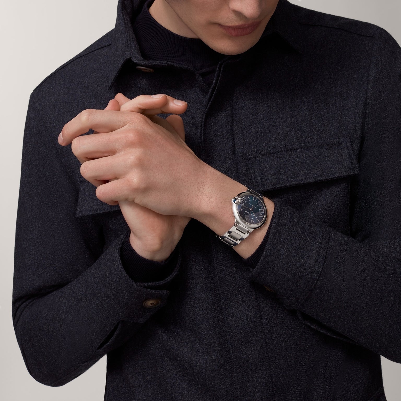A person in a dark jacket wears a Cartier Ballon Bleu de Cartier watch, its black face and steel bracelet visible as their hands are clasped together near their chest.