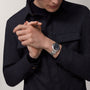 A person in a dark jacket wears a Cartier Ballon Bleu de Cartier watch, its black face and steel bracelet visible as their hands are clasped together near their chest.
