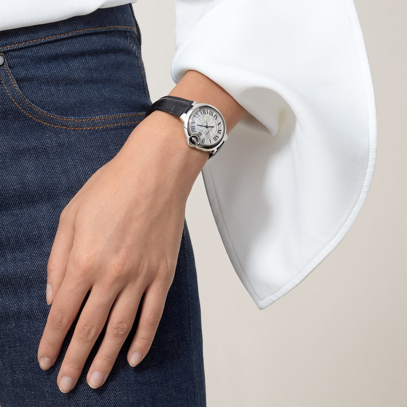 A person wearing blue jeans, a white long-sleeve top, and a Ballon Bleu de Cartier watch with an alligator leather strap by Cartier on their left wrist.