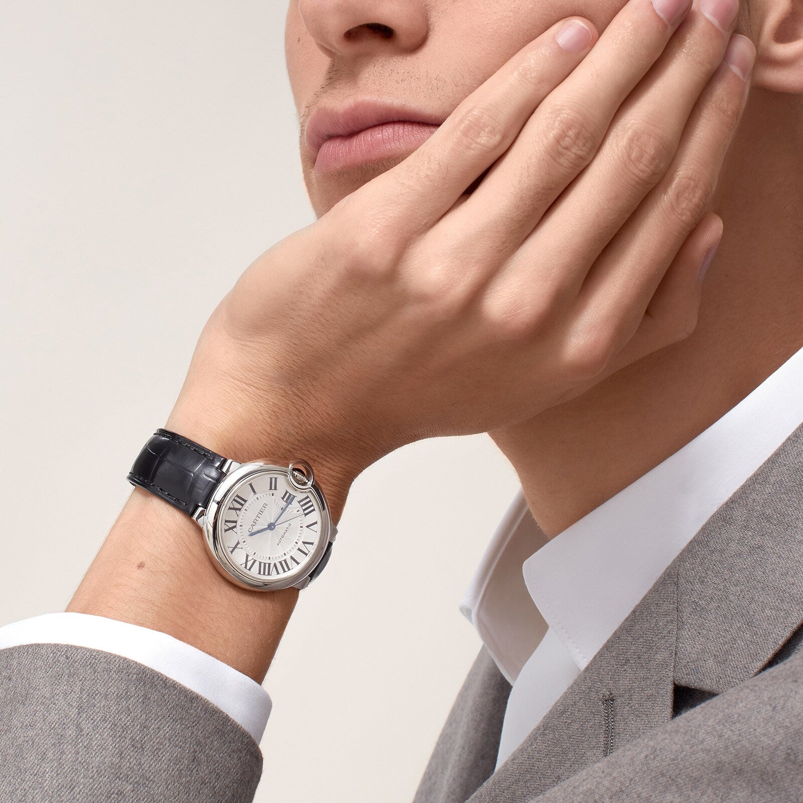A person in a gray suit rests their chin on their hand, showcasing a Cartier Ballon Bleu de Cartier watch with a white face and black alligator leather strap.