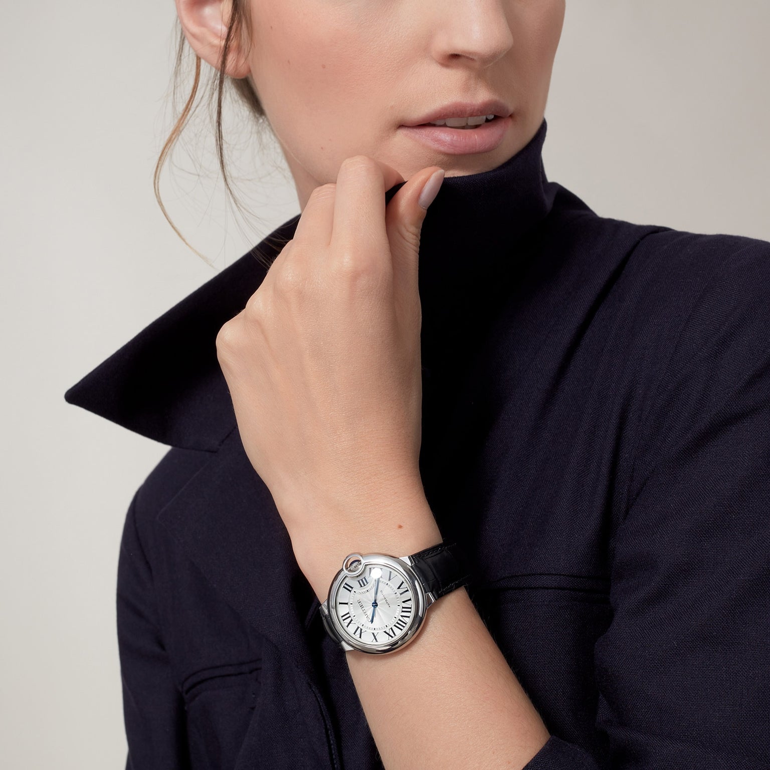 A person in a dark blazer holds their collar, showcasing the Cartier Ballon Bleu de Cartier with Roman numerals, a round silver case, and a black alligator leather strap.