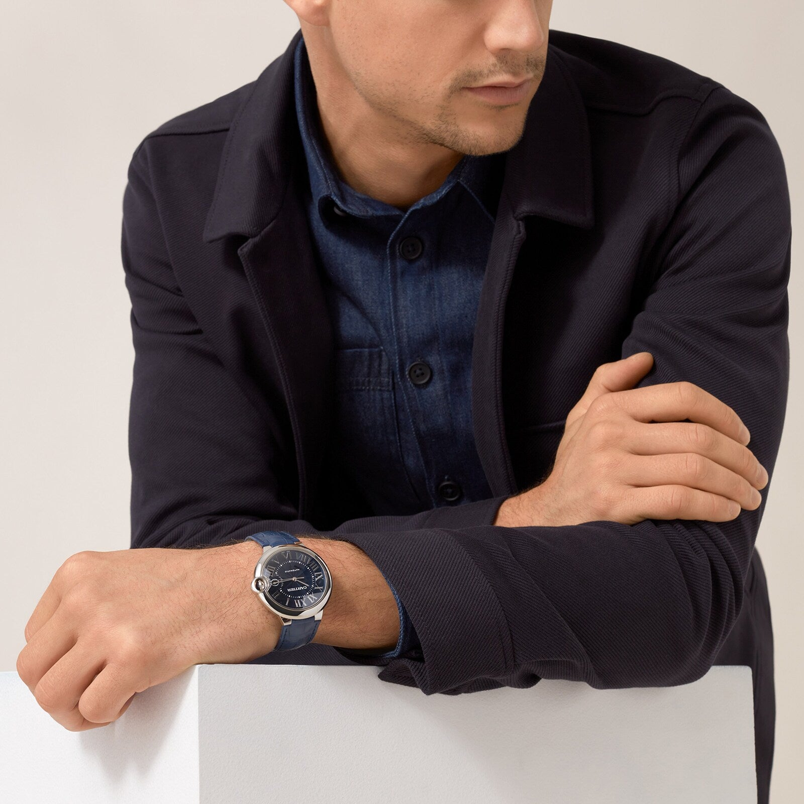 A man in a dark jacket and denim shirt rests his arms on a white surface, wearing the Cartier Ballon Bleu de Cartier with a striking blue guilloché dial on his left wrist.