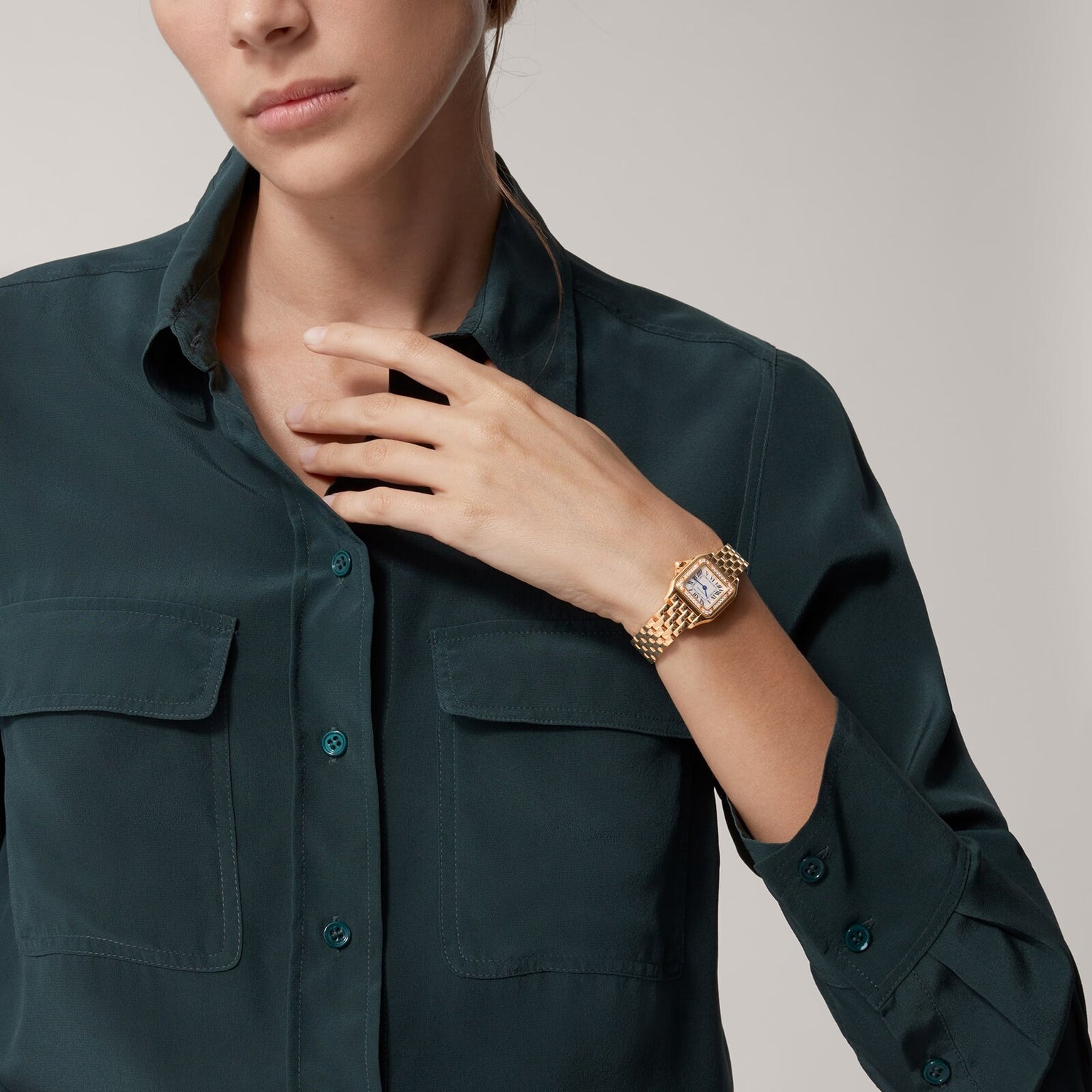 A woman in a dark green button-up shirt showcases a Cartier Panthère de Cartier Small Model gold wristwatch on her left wrist, her hand poised near her collar.