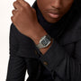 A person in a dark coat wears a Cartier Santos de Cartier silver wristwatch with a black dial, metal bracelet, and automatic self-winding movement on their left wrist.