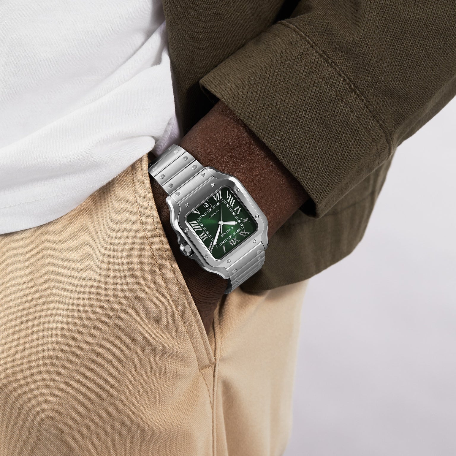 A person displays form precision wearing Cartier’s Santos de Cartier watch with a silver case, green dial, and Roman numerals, styled with a white shirt, olive jacket, and beige pants, one hand tucked into their pocket.