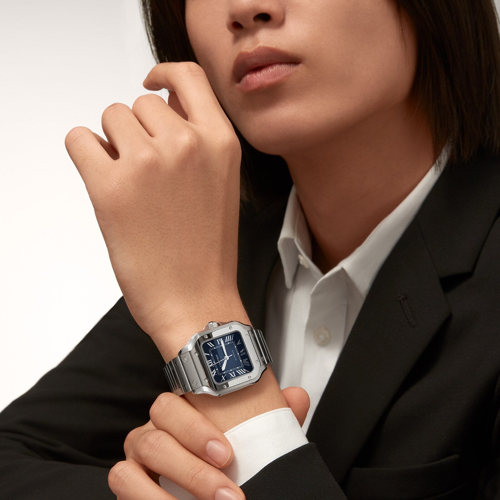 A person in a black blazer and white shirt showcases a Cartier Santos de Cartier wristwatch with a blue square face and square steel case on their left wrist.