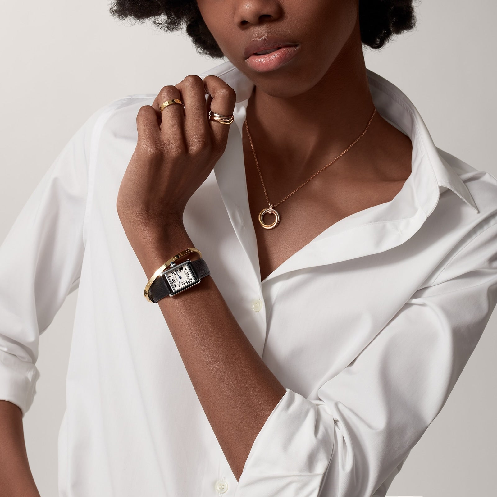 A person wearing a white button-up shirt, gold jewelry, and a square-faced Cartier Tank Must de Cartier watch with quartz movement poses with one hand near their neck.