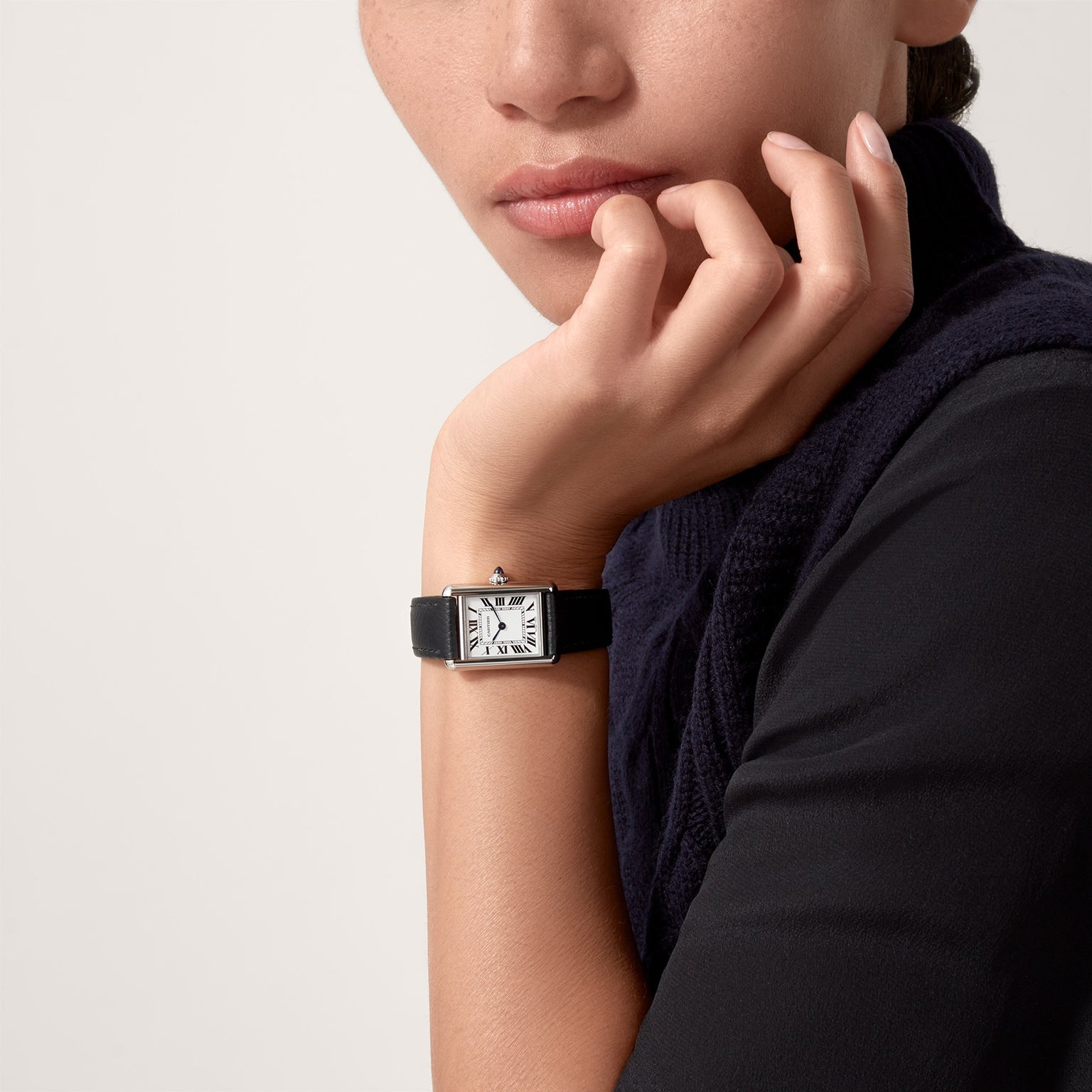A person in a dark sweater rests their chin on their hand, showcasing the Cartier Tank Must de Cartier watch featuring a beaded crown and Roman numeral dial on a black leather strap.