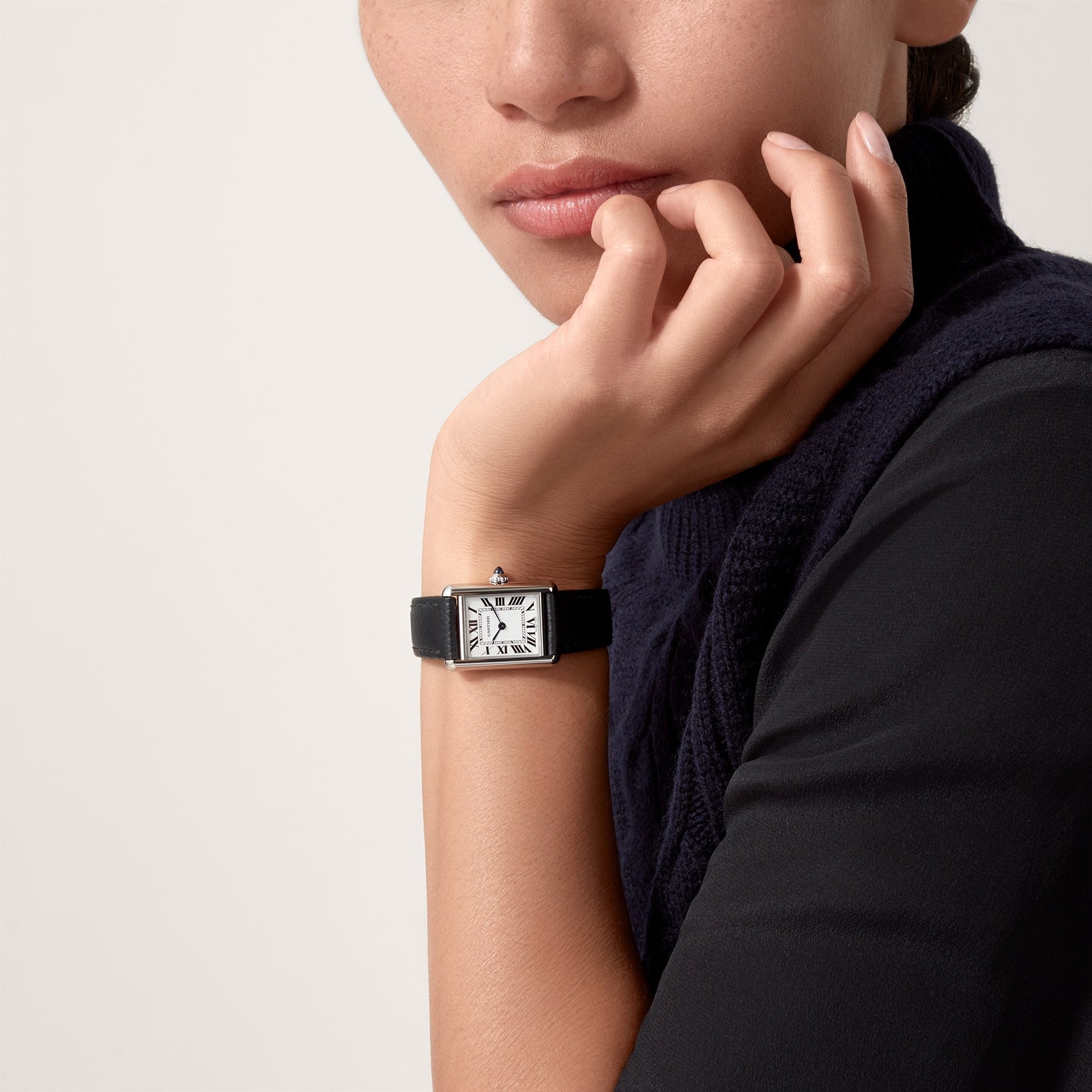 A person in a dark sweater rests their chin on their hand, showcasing the Cartier Tank Must de Cartier watch featuring a beaded crown and Roman numeral dial on a black leather strap.