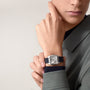 Someone in a gray coat displays their wrist adorned with a Cartier Santos-Dumont watch, featuring a silver square case, Roman numeral dial, and black leather strap.