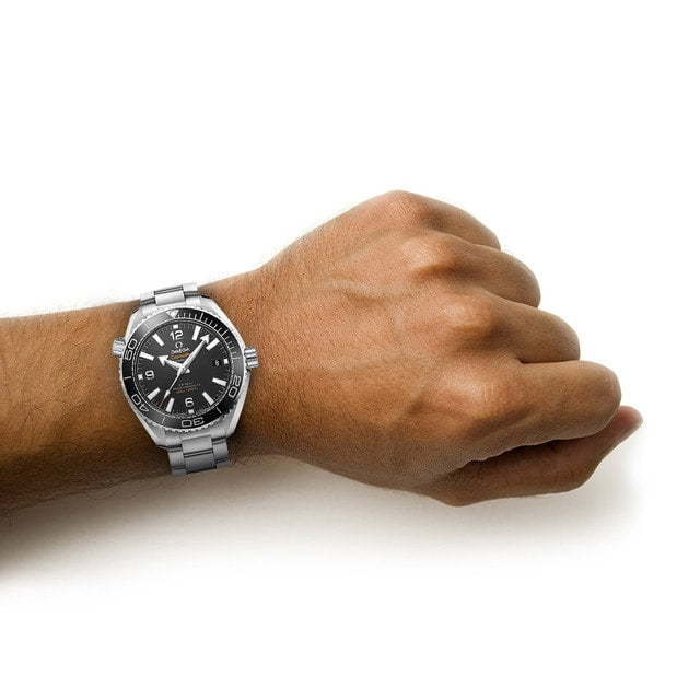 A persons left wrist wearing an OMEGA Seamaster Planet Ocean 600M diving watch with a black face and silver band, set against a white background.