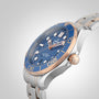 A close-up of the OMEGA Seamaster Diver 300M wristwatch, featuring a blue face, gold accents, and a silver-gold metal band, inspired by James Bond, set against a light gradient background.