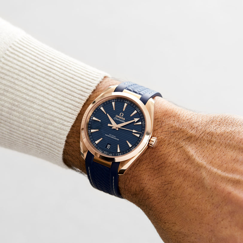 An individual wears an OMEGA Seamaster Aqua Terra 150M with a gold case, blue dial, and blue strap on their left wrist, the watch partly visible under a light-colored sweater sleeve.