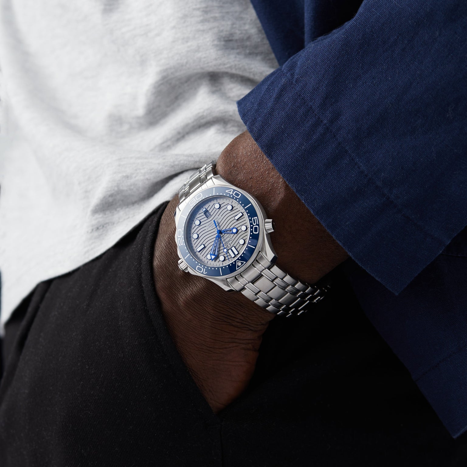 A person wearing an OMEGA Seamaster Diver 300M with a blue dial and silver band, dressed in a white shirt, navy jacket, and black pants, stands with one hand in their pocket.