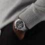 A person in a grey sweater and black pants has their hand in their pocket, showcasing an OMEGA Seamaster Diver 300M watch with a sleek black dial.