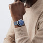 A person in a beige sweater rests their chin on their hand, showcasing the OMEGA Constellation wristwatch with a blue face and an eye-catching blue ceramic bezel.