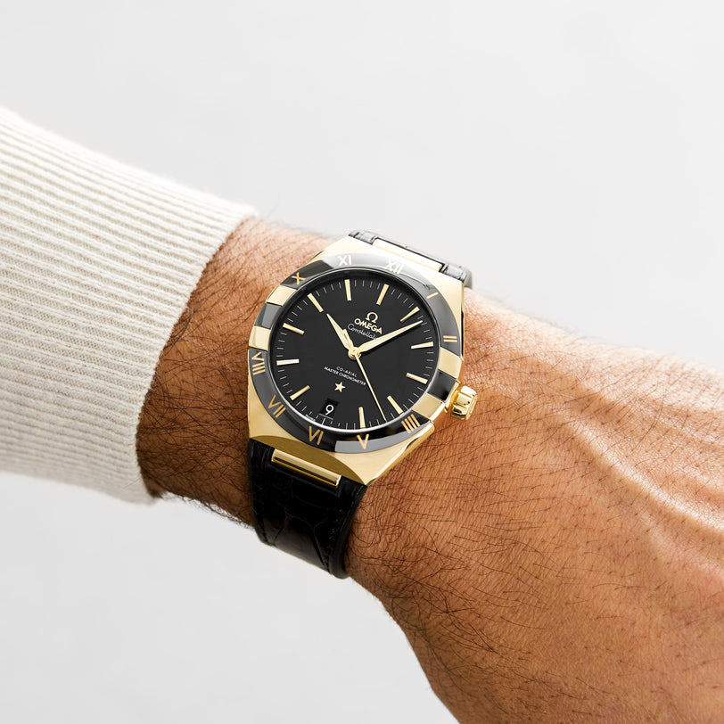 A wrist wears an OMEGA Constellation watch in gold and black with Roman numerals on a black leather strap, showcased against a plain background.
