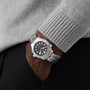 A person in a gray sweater and black pants has their hand in their pocket, showcasing the OMEGA Seamaster Diver 300M Nekton Edition, featuring a laser-ablated ceramic dial and stainless steel case.